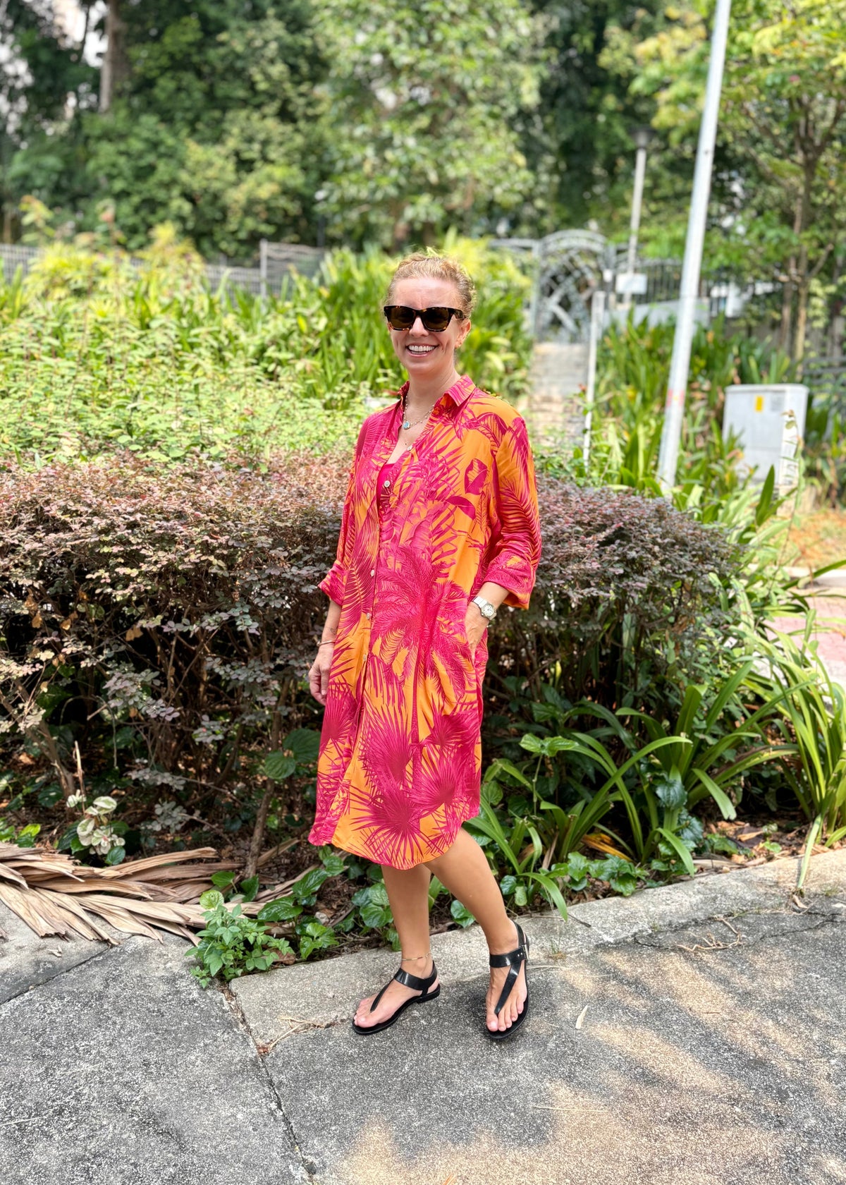 A woman wearing a vibrant pink and orange tropical floral shirt dress with pockets, styled with black sunglasses and black sandals, standing outdoors on a paved path with green bushes in the background.
